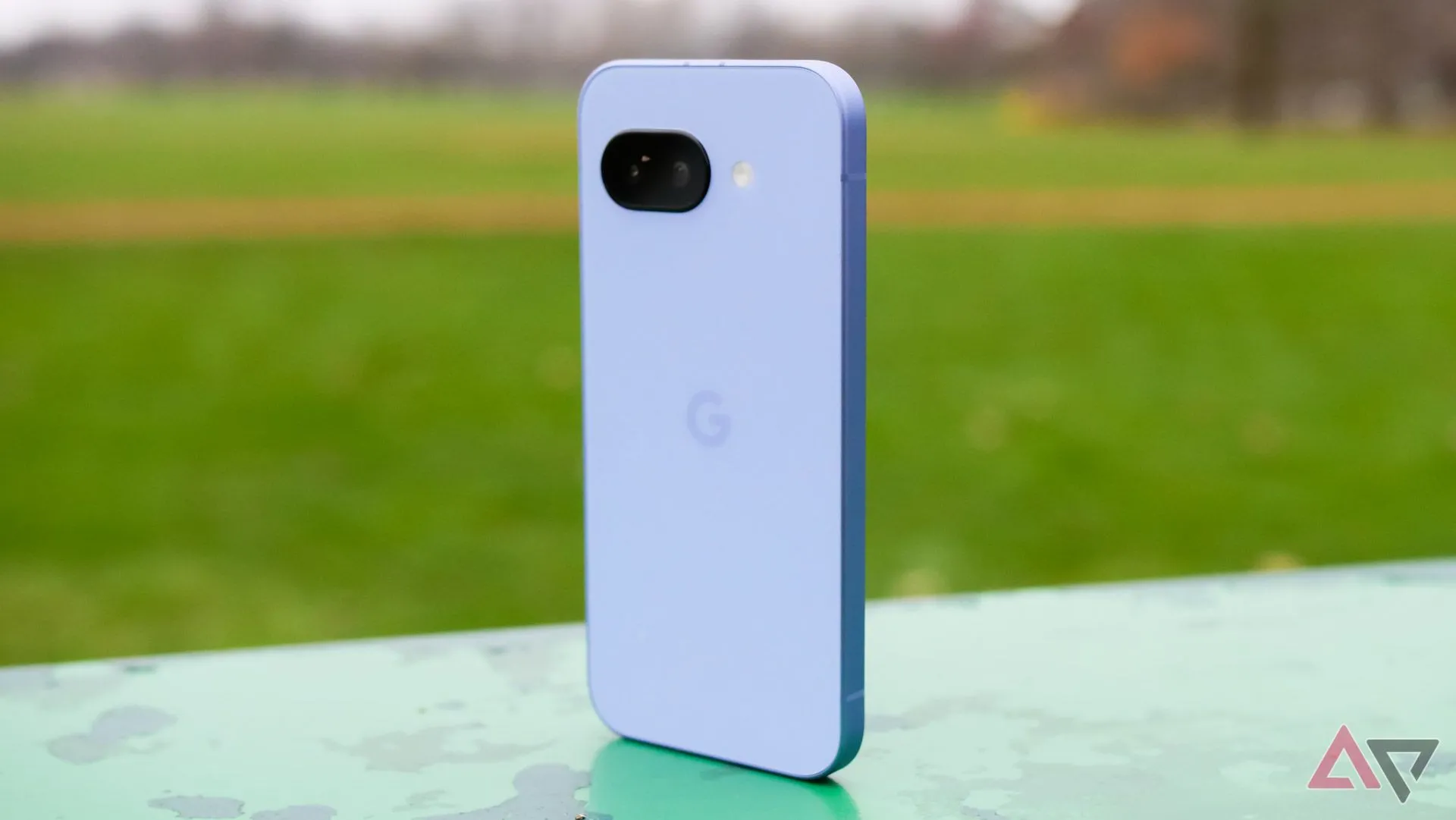 Điện thoại Google Pixel 9a màu tím đặt đứng trong công viên, mặt lưng hướng về phía trước.
